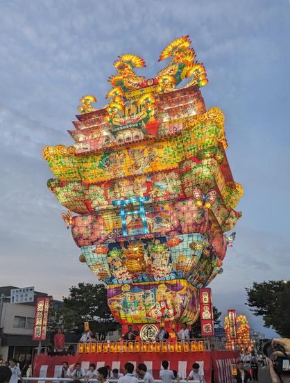 Large, illuminated lantern float being paraded for a japanese summer festival. It is made of colorful paper and lamps and appears to be pulled by a group of people.