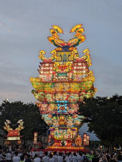 Large, illuminated lantern float being paraded for a japanese summer festival. It is made of colorful paper and lamps and appears to be pulled by a group of people.