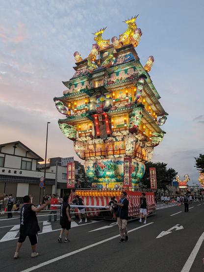 Large, illuminated lantern float being paraded for a japanese summer festival. It is made of colorful paper and lamps and appears to be pulled by a group of people.