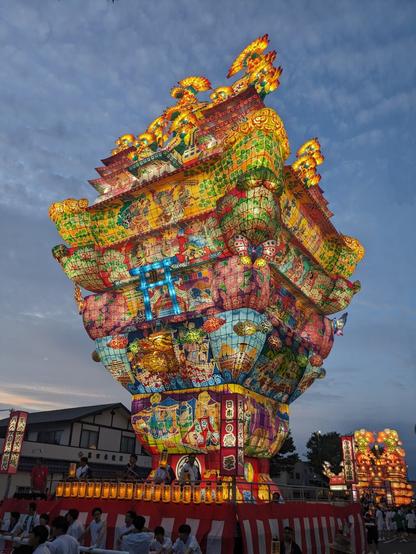 Large, illuminated lantern float being paraded for a japanese summer festival. It is made of colorful paper and lamps and appears to be pulled by a group of people.