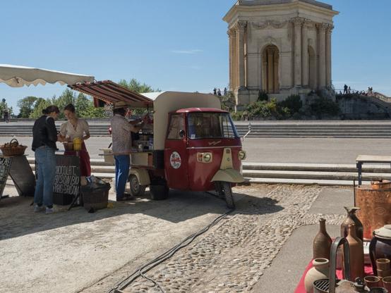 The image captures a picturesque scene in Montpellier, France. In the foreground, a converted red Piaggio Ape serves as a mobile coffee and snack stand. Beneath a striped awning, organic treats and beverages are offered, while guests engage in a relaxed interaction, adding to the lively atmosphere.

In the background stands the impressive Château d'Eau in the Jardin du Peyrou, a well-known landmark of Montpellier. This elegant neoclassical structure, with its columns and arches, was historically part of a water reservoir and remains a popular destination for locals and tourists alike. The sunny weather, historical charm, and rustic details combine to create a vibrant and inviting ambiance.