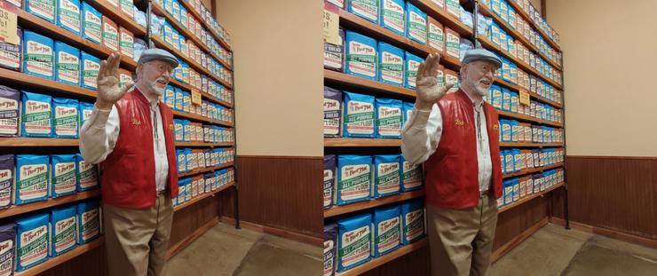 Stereoscopic, side-by-side view. A sculpture of Bob Moore, of Bob's Red Mill, standing indoors in front of a wall of packages of flour. An older gentleman with a beard and wire-framed eyeglasses. He is waving his right hand, and is wearing his trademark pale blue flat cap, string tie, red vest and khaki trousers. (The statue is painted in color)