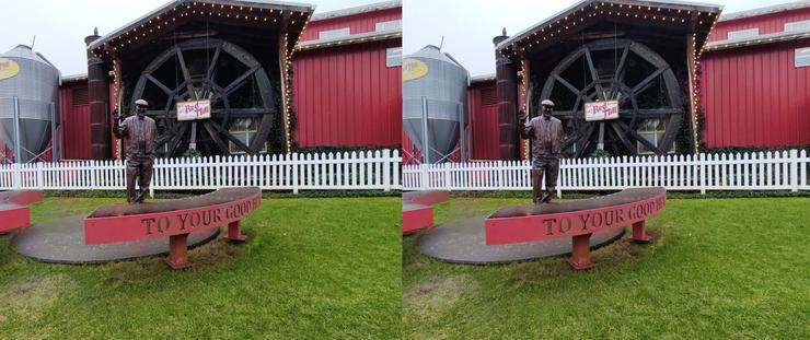 Stereoscopic, side-by-side view. A bronze sculpture of Bob Moore, of Bob's Red Mill, standing on a lawn outdoors between a pair of semi-circular benches, in front of a waterwheel incorporated into a red building. He is waving his right hand, and is wearing his trademark flat cap, string tie and vest.
