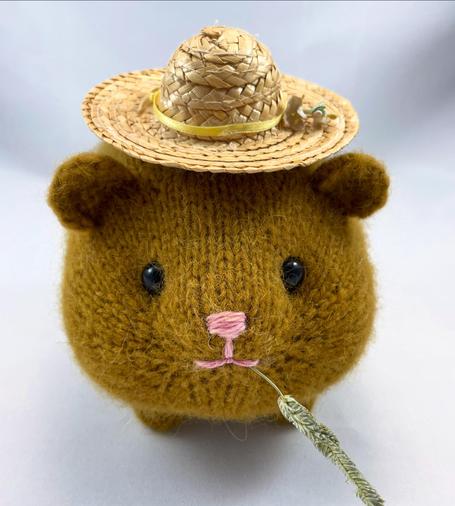 photo of a knitted golden brown guinea pig, wearing a little straw hat, and nibbling on a piece of hay