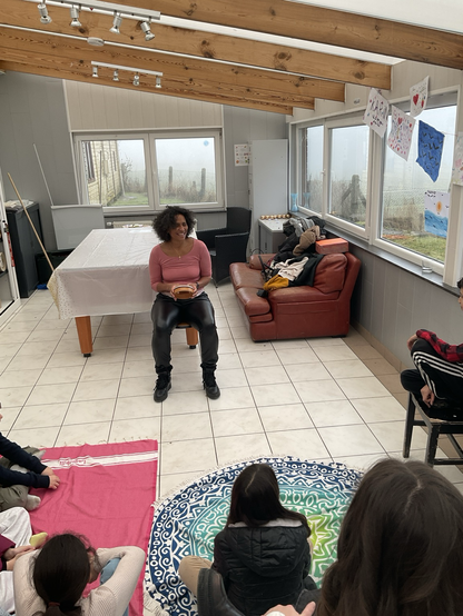 Ria Carbonez, conteuse, assise sur un tabouret dans une véranda lumineuse, tenant un petit tambour. Autour d’elle, des enfants et adolescents sont assis sur des tapis colorés, écoutant attentivement. La pièce est décorée de dessins accrochés aux fenêtres et d’un canapé en cuir rouge