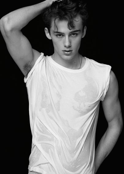 Greyscale photo of model William Franklyn-Miller, upperbody visible, with short curly hair in a wet white t-shirt, posing with one hand behind their head against a dark background.