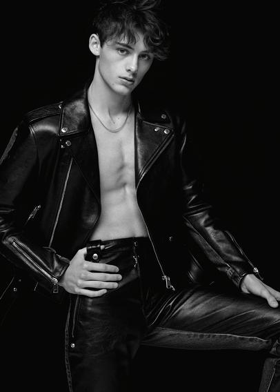 Greyscale photo of model William Franklyn-Miller, upperbody and upper legs visible, with short curly hair in an opened leather jacket, posing with hands placed on his pants against a dark background.