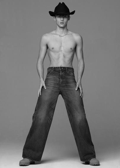 Greyscale photo of model William Franklyn-Miller,whole body visible, shirtless with a cowboy hat and jeans, posing with hands placed on his pants against a light background.