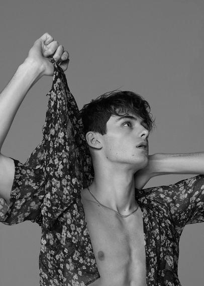 Greyscale photo of model William Franklyn-Miller,upperbody visible, lifting arm, wearing an opened floral-patterned garment, looking to the side.