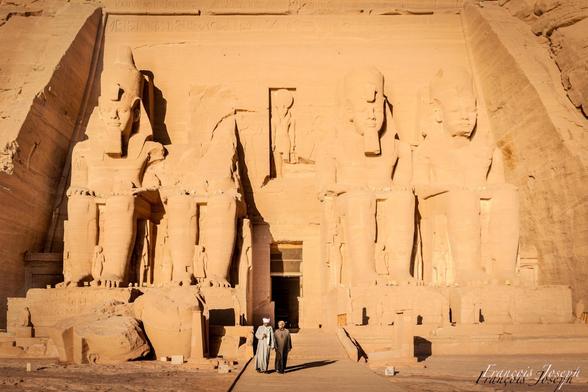 🇫🇷
“Photographie du temple d’Abu Simbel, en Égypte, au lever du soleil. La façade monumentale sculptée dans la roche représente quatre colosses assis de Ramsès II, dominant l’entrée du temple. Deux hommes en djellaba avancent paisiblement sur le chemin menant à l’entrée, offrant une échelle humaine à la grandeur du site. La lumière dorée du matin éclaire les statues, mettant en valeur les reliefs et les hiéroglyphes gravés sur les parois.”

🇬🇧
“Photograph of the Abu Simbel temple in Egypt at sunrise. The monumental rock-carved façade features four seated colossi of Ramses II towering over the temple entrance. Two men in djellabas walk peacefully along the path leading to the doorway, providing a human scale to the site’s grandeur. The golden morning light illuminates the statues, highlighting the relief carvings and hieroglyphs etched into the walls.”