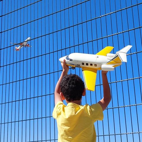 Criança com camisola amarela segura no ar um avião amarelo de brincar, enquanto ao fundo um avião verdadeiro aparece