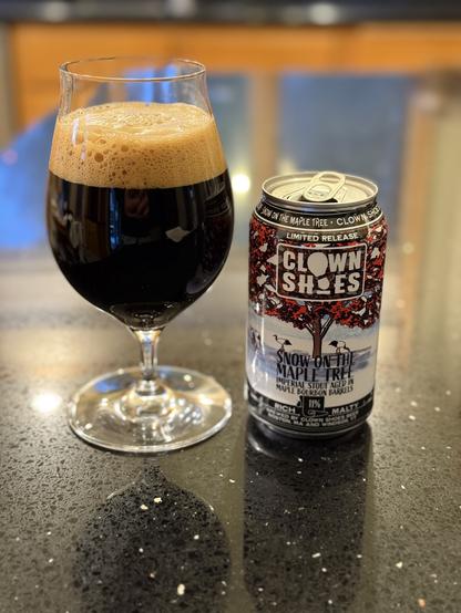 A snifter glass of dark beer with a tan head. The 12 ounce can to the right has a red-leaved Maple tree on a snowy white background