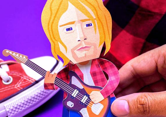 Paper cutout figure of Kurt Cobain holding a guitar. A hand adjusts the figure near a red shoe against a purple background.