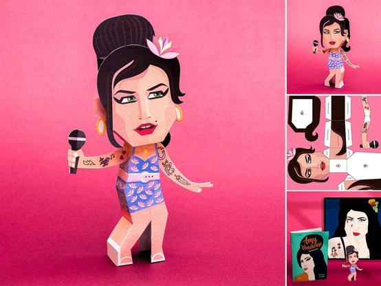 Paper doll of Amy Winehouse with vintage hairstyle, tattoos, and microphone. Side images show assembly instructions and packaging on a pink background.