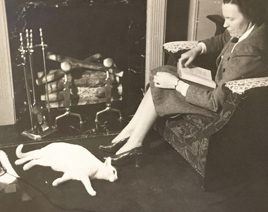 Black and white photo of a white woman in smart blazer and matching skirt, reading in an armchair by a fireplace. A shorthaired white cat is lying on the floor in front of the unlit fireplace, sprawled out looking bored.