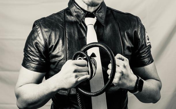 A black and white photograph represents the leather fetish aesthetics. The composition focuses on a torso wearing a sleek black leather uniform shirt with short sleeves, creating a contrast against a light background. A crisp white tie adds a formal, commanding element to the ensemble. The central focus is strong hands gripping a braided leather signal whip, held in a purposeful stance that suggests both control and expertise. A watch adds a modern touch to this classic fetish imagery. The image is cropped to maintain anonymity while emphasizing the visual elements of leather, discipline, and authority. The high-contrast monochrome treatment enhances the visual impact of the textures and forms.