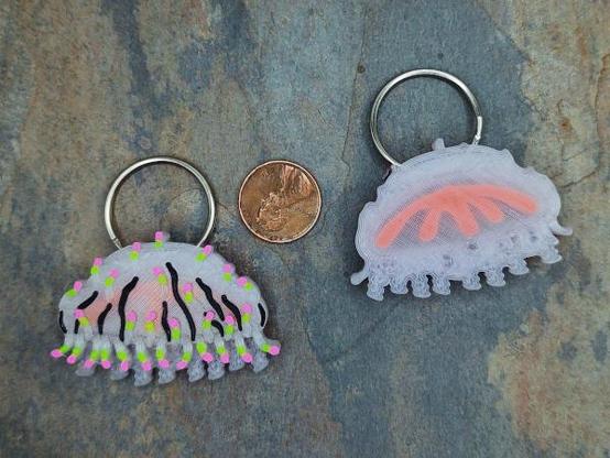 Two 3d printed Flower-hat Jelly designs. On the left, a jelly-fish like creature with translucent bell and coiled tentacles. The bell has vertical black stripes and short tentacles tipped with neon green and pink. The left design shows the “back” of the design, with an orange inner structure visible through the translucent body. Both designs have-inch (2.5cm) split ring through the top of the bell. In the middle is a US penny for scale.