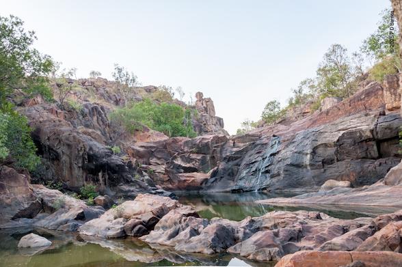 Looking up-hill at the creek-bed of a seasonal high-rainfall waterfall creek running through a solid sandstone escarpment. There is currently very little water flowing, but the width of the bed and the degree of erosion clearly indicates that very large volumes of water run down in the wet season. The view shows a pool with large exposed sandstone rocks worn smooth by water, while further upstream is a rock wall several metres high, also worn in the centre, which blocks the view of the creek. Rough sandstone walls rise up on either side of the creek bed.
