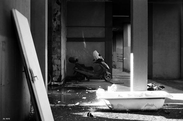 Schwarzweißfoto, aufgenommen vor einem heruntergekommenen Wohngebäude mit bequem entsorgten Objekten, darunter einer angelehnten Tür links, einer Badewanne unten rechts und ein Motorroller hinten in der Mitte. Leica R5 mit Summicron 35 mm auf Agfa APX 100-Film.