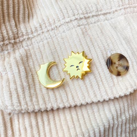 A photo of two mini pins stuck to the pocket of a curderoy shirt. The pins are of a sun with a cat face and a crescent moon with cat ears.