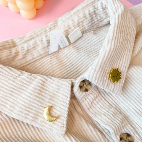 A photo of two collar pins stuck to the collar of a corduroy shirt. The pins depict an adorable golden sun with a cat face and a golden crescent moon with cat ears.