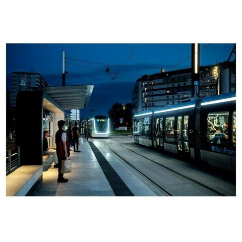 Une station de tramway en Île-de-France est capturée à la tombée de la nuit, baignée dans une lumière artificielle qui contraste avec le bleu profond du ciel. Un tramway est arrêté sur la droite, ses vitres reflétant la lumière et laissant entrevoir des passagers assis à l’intérieur. Un autre tram approche au loin, ses phares blancs dessinant une lueur futuriste sur les rails.

Sur le quai, plusieurs personnes attendent, chacune plongée dans son univers. Un homme debout, un sac en papier marqué “Jules” à la main, regarde en direction du tram. Un autre, assis sous l’abri moderne aux lignes épurées, est absorbé par son téléphone. Plus loin, une femme portant un sac blanc avance vers le tram en approche.

En arrière-plan, de grands immeubles de logements aux fenêtres illuminées composent le décor urbain. Les câbles électriques du tramway se détachent contre le ciel nocturne, ajoutant une dynamique graphique à la scène. L’image évoque à la fois l’agitation discrète du quotidien et une atmosphère presque cinématographique, portée par les lumières et les reflets.
