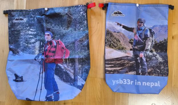 Two dry bags, the left being slightly larger than the right. The left bag has an image on a ski mountaineer standing on skis with a red backpack. The right bag has an image of a hiker with a finger pointing to the top of Mt Everest. The words ysb33r in Nepal is printed on the bottom.