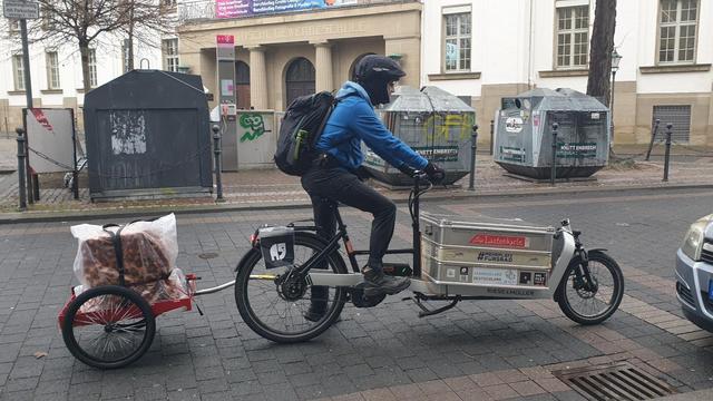 Blick vom Gehweg auf einen Radfahrer, der auf einem Long-John-Lastenrad mit Box sitzt. Am Lastenrad hängt ein Anhänger. Auf dem Anhänger ist ein Farblaser-Multifunktionsdrucker in eine Decke und einen Kunststoffsack gehüllt und verzurrt.