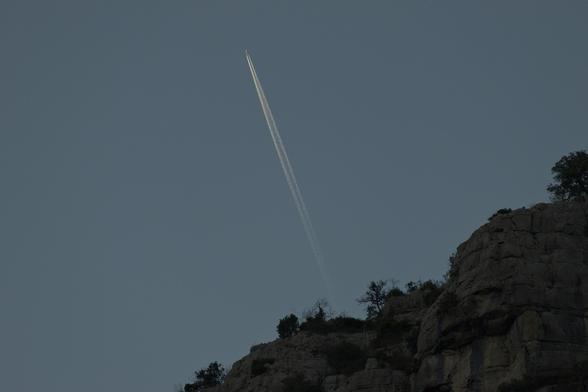 Un avion et sa trace blanche dans un ciel dégagé du crépuscule. Au-dessous, on distingue la forme de falaises déjà dans l'obscurité.