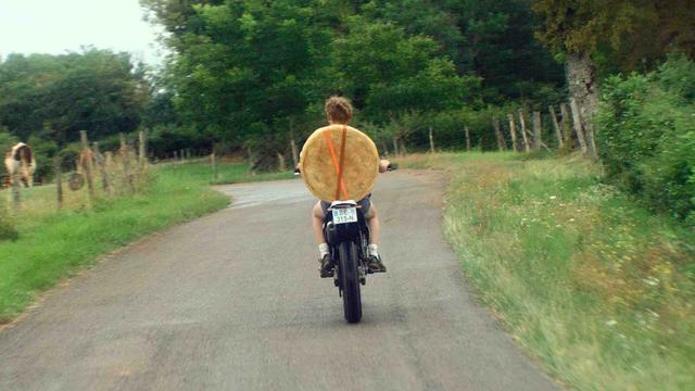 Ein junger Mopedfahrer fährt durch eine ländliche Gegend. Auf seinen Rücken hat er sich ein großes Käserad geschnallt.