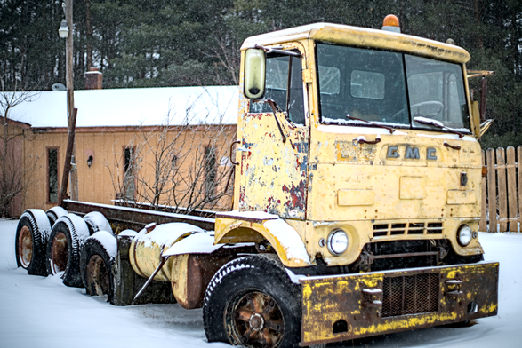 A weathered yellow GMC truck with a cab-over design, missing its bed, sits abandoned on a snowy roadside. The vehicle shows significant rust and paint chipping, with snow accumulating on its tires and frame. Behind the truck, a wooden building and a tall wooden fence are visible, surrounded by trees and falling snow.
