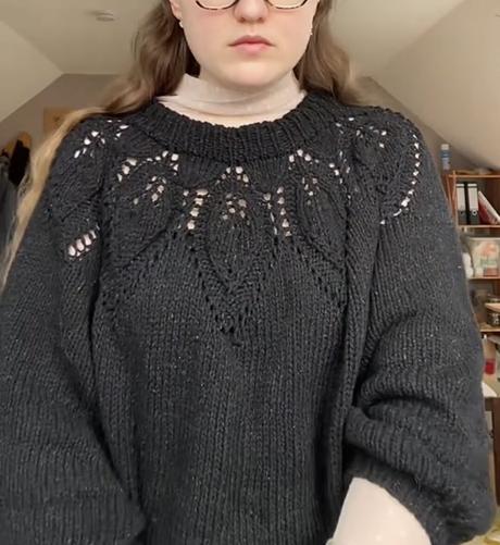 Mauni a white person is wearing a black hand knitted sweater with a leaf pattern at the collar