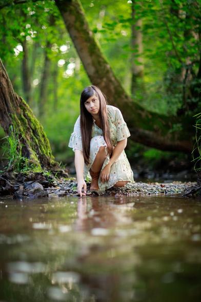 A young woman with long dark hair, wearing a light floral dress and brown boots, is crouched by the edge of a forest stream. She is gently touching the water with her fingers, surrounded by lush green trees and moss-covered roots, creating a serene and peaceful natural scene.