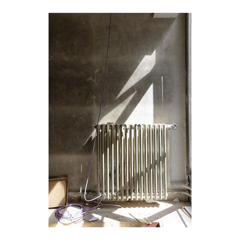 Un radiateur en fonte couleur beige devant un mur poussiéreux. La lumière d'une fenêtre découpe des formes géométriques sur les objets.