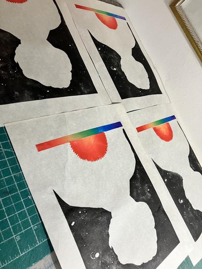5 of my prints are laid out drying on a tabletop, now in their third state. In addition to the thin band along (what will be) the bottom with a left to right rainbow spectrum of violet through to red, just above that in the middle is a semicircular shape with radiating spikes printed in a radial gradient of yellow in the centre out to red. Also, around what is currently an unprinted silhouette of a short haired woman from the chest up, facing forward. Is a background in black like space with carved stars and galaxies. The prints are awaiting their final layer with my portrait of Burbidge, lined and labels within the semicircle of supergiant star and stellar absorption lines along the  spectrum at the bottom.