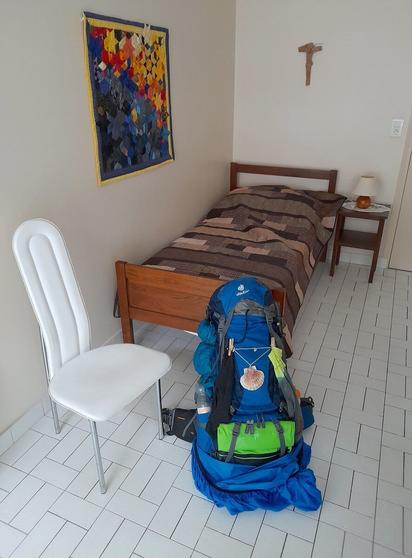 A sober, somewhat old-fashioned looking bedroom. A large, bright blue backpack is leaning against the bed. To the left is a colourful quilt on the wall and above the headboard of the bed a cross is hanging.