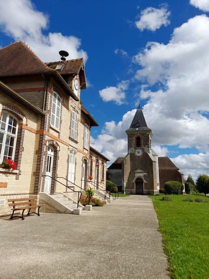 A sunny day with a bright blue sky. To the left is a French townhall with a bench in front of it and ahead, further in the distance, is a small church.
