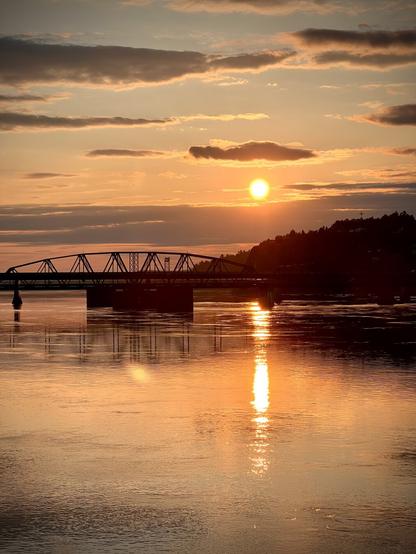 This image captures a beautiful sunset over a body of water, with the sun low on the horizon casting a warm golden glow. The reflection of the sun and clouds can be seen on the water's surface, creating a serene and picturesque scene. A bridge is silhouetted against the sunset, adding to the composition. The overall atmosphere is tranquil and inviting.