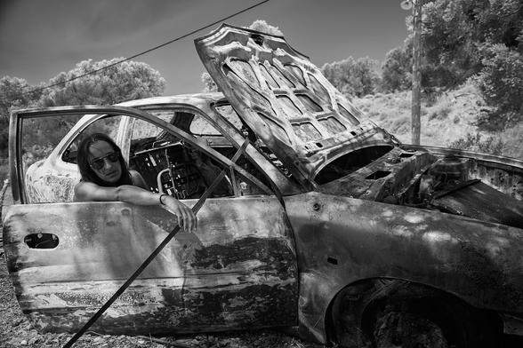 A black and white picture of a burnt out car wreckage in a rural landscape. The right door is open and a woman with sunglasses is leaning her right arm out of the window.