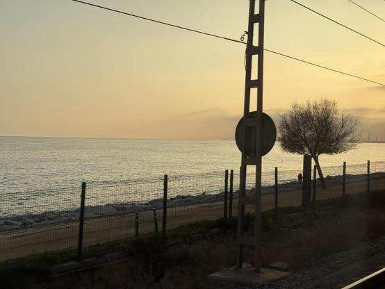 Foto hecha desde la ventana de un tren, se ve el atardecer con pocas nubes, parte de la vía de al lado y un poste de la catenaria con la parte trasera de una señal.

Además hay una valla, detrás de ésta un camino de tierra con un árbol en la derecha y unas piedras para contener el oleaje.

De lejos se ve parte de las Tres Xemeneies de la antigua central eléctrica de Sant Adrià de Besòs.