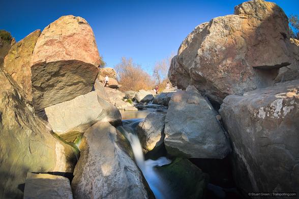 A scenic view of a small waterfall flowing through large boulders in a rocky landscape. The water cascades down between the rocks, creating a smooth, silky effect. The sky is clear and blue, and there are a few people in the background, standing on the rocks and observing the scene.