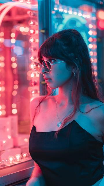 🇫🇷  Une jeune femme avec un top noir et des lunettes de soleil, posant devant une attraction lumineuse de fête foraine. Les lumières rouge et bleu enveloppante donne une ambiance année 80.

🇬🇧 A young woman in a black top and sunglasses, posing in front of a fairground attraction. The enveloping red and blue lights give it an 80s feel.