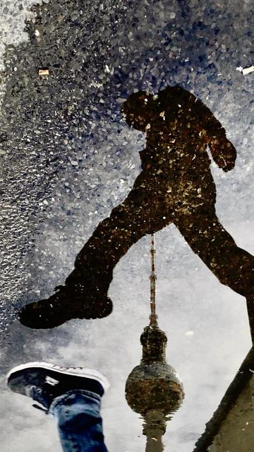A reflection in a puddle shows a silhouette of a person walking, with one foot visible in the foreground. The reflection also includes a tower, appearing to be a landmark, beneath the figure. The ground is wet and textured, enhancing the visual contrast.