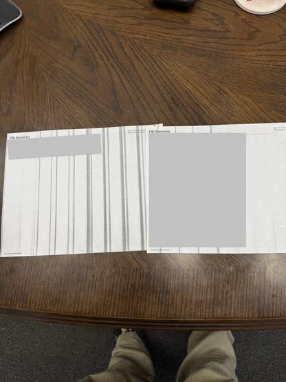The image shows a wooden table with a document laid out on it. The document appears to be an "F&I Summary" and is printed on two pages. Most of the content on the pages is obscured by gray rectangles, possibly for privacy reasons. At the top right corner of each page, it says "Date: Thu Feb 16, 2023" and "Report Period: 02/16/23". At the bottom of each page, it says "Powered by StoneEagle". In the background, part of a computer mouse and a coaster with a drink are visible on the table. The lower part of the image shows a person's legs and feet, wearing khaki pants and dark shoes, standing in front of the table.
