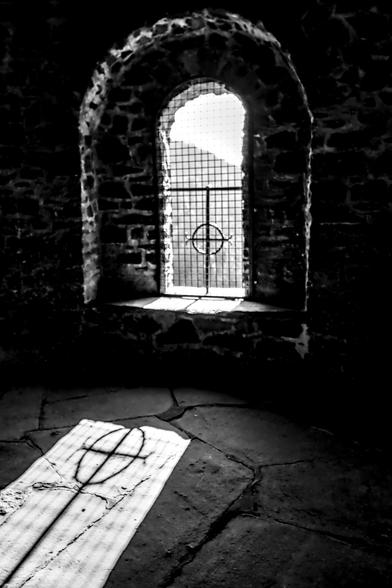Das Bild zeigt einen Innenraum mit einem vergitterten Fenster in einer Steinmauer. Das Fenster wirft einen Schatten mit einem Kreuzsymbol auf den Steinboden. Das Bild ist in Schwarz-Weiß gehalten