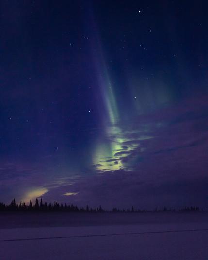 In the lower part of the image, the frozen Muonio River is visible. On the opposite shore, on the Swedish side, there is a dimly lit forest. The sky is filled with stars and the northern lights, with clouds drifting in front of them.