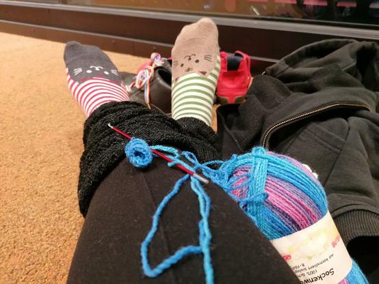 The legs and feet of a human can be seen sitting on a beige carpet. The legs, clad in black leggings and black gaiters, are crossed, with a red and white curled sock with a black cat face on one foot and a green and white curled sock with a beige cat face on the other foot. The cat faces are in the toe area of each sock. In front of the feet is a pair of black shoes with rainbow shoe laces and a single red cotton barefoot shoe. A sloppily thrown black hooded jacket to the right of the thighs. A twisted crocheted curl in blue crocheted onto a metallic red crochet hook. The wool thread comes from a colour gradient ball of wool with a colour spectrum ranging from pink to purple to blue. Half of the text on the label reads: 'Sock wool, 100% virgin wool from certified organic farming, 8-ply'. It actually reads this: 'Sockenwo - 100% Schu - aus kontrolliert biologi  - 8-fädi'