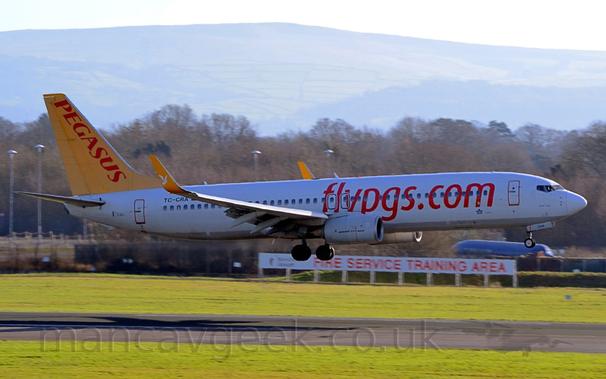 Side view of a white, twin engined jet airliner with red billboard-style "FlyPGS[dot]com" titles on the forward fuselage, yellow upturned wingtips with white winged horses in the middle, and a yellow tail with red "Pegasus" titles.
The plane is flying from left to right at a very low altitude, about to land on the black runway.
Green grass fills the foreground, either side of the runway, with a white sign with red text "Fire training area" in front of a black fence.
The blue fuselage of a plane and trees fill the rest of the background, under grey skies.