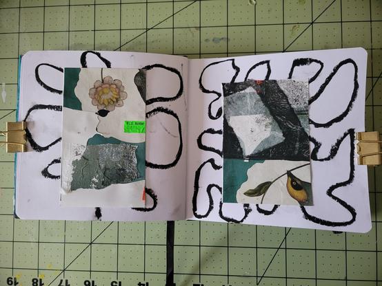 An open sketchbook on a gridded cutting mat, held open with gold binder clips. The pages feature abstract mixed-media collages with torn paper, floral and bird imagery, and a bold black oil pastel design behind resembling loops or petals