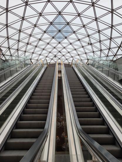 A pair of long, parallel escalators ascend towards a vanishing point beneath a striking geometric glass dome at Triangeln Station in Malmö, Sweden. The dome's intricate metal framework forms a repeating triangular pattern, allowing natural light to filter through. Glass railings on either side reflect the escalators, enhancing the sense of depth and symmetry. Beneath the escalators, a glimpse of warm lighting and spherical chandeliers contrasts with the station’s sleek, modern aesthetic. The perspective creates a dynamic, almost futuristic feel, drawing the eye upward towards the light-filled exit